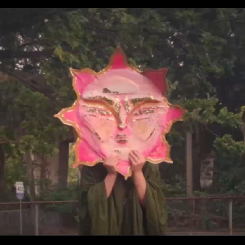 A pink and gold sun mask being held up to cover the models face. They are outside in a park.