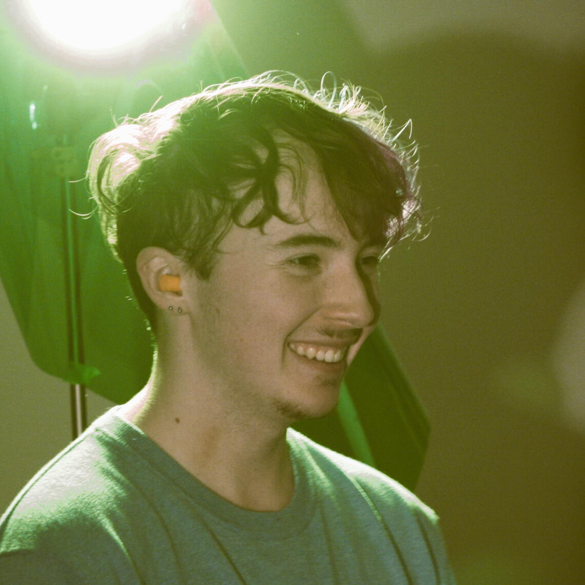 A side profile photo of a young man with a goatee and blue shirt smiling. The photo is tinted green.